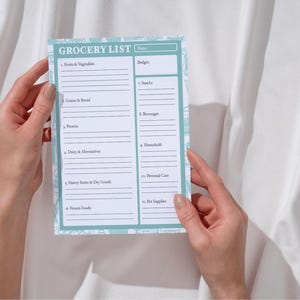 Pode incluir: Um bloco de notas de lista de compras com uma borda azul clara e detalhes florais é segurado por duas mãos. O bloco de notas tem seções para frutas e vegetais, grãos e pão, proteínas, laticínios e alternativas, itens de despensa e produtos secos, etc.