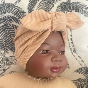 May include: A baby wearing a light brown turban with a bow on top. The turban is made of a soft, ribbed fabric.