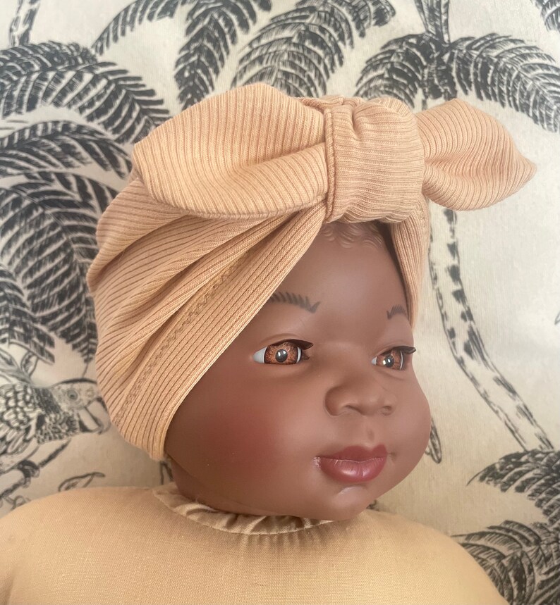 May include: A baby wearing a light brown turban with a bow on top. The turban is made of a soft, ribbed fabric.