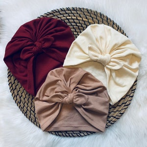 May include: Three turban headbands in different colors: burgundy, beige, and cream. Each headband has a large bow on top.