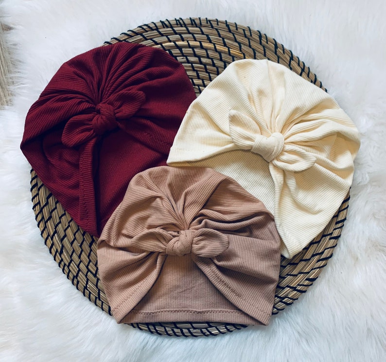 May include: Three turban headbands in different colors: burgundy, beige, and cream. Each headband has a large bow on top.