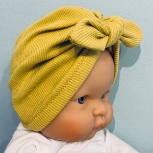 May include: A yellow ribbed turban with a bow on top. The turban is made of a soft, stretchy fabric and is perfect for keeping your hair out of your face.