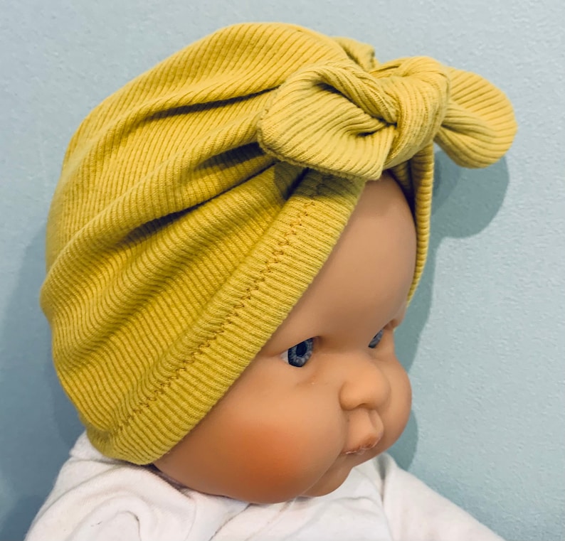 May include: A yellow ribbed turban with a bow on top. The turban is made of a soft, stretchy fabric and is perfect for keeping your hair out of your face.