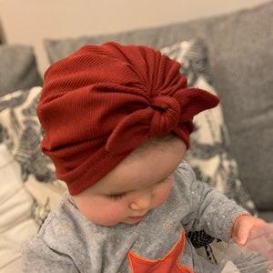 May include: A baby wearing a red ribbed turban with a bow on top. The baby is wearing a gray shirt with a red star on it.