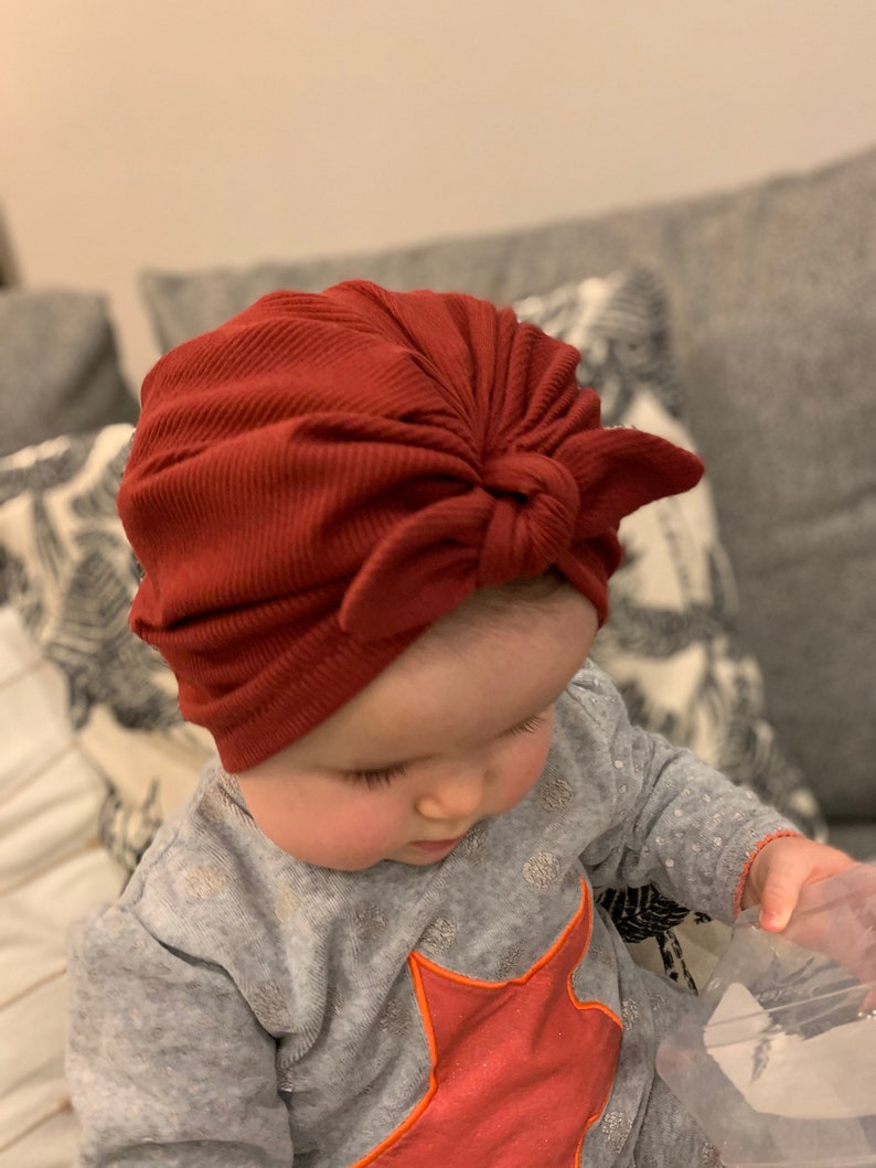May include: A baby wearing a red ribbed turban with a bow on top. The baby is wearing a gray shirt with a red star on it.