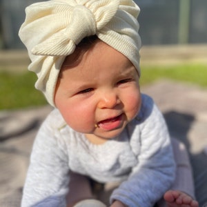 May include: A baby wearing a white turban with a bow on top, sitting on a gray blanket and holding a clear baby bottle.