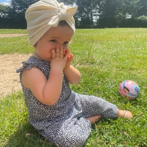 May include: A young child wearing a white turban and a black and white floral jumpsuit sits on the grass. The child is looking at the camera and has their hands covering their mouth. A pink and blue ball is in the background.