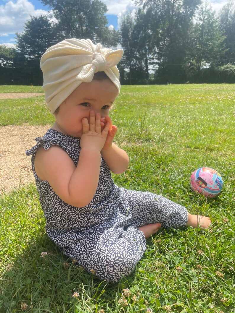 May include: A young child wearing a white turban and a black and white floral jumpsuit sits on the grass. The child is looking at the camera and has their hands covering their mouth. A pink and blue ball is in the background.
