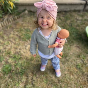 May include: A young child wearing a pink turban with a bow on top is holding a doll. The child is smiling and looking at the camera. The child is wearing a light green cardigan and blue jeans. The child is standing on a grassy surface.