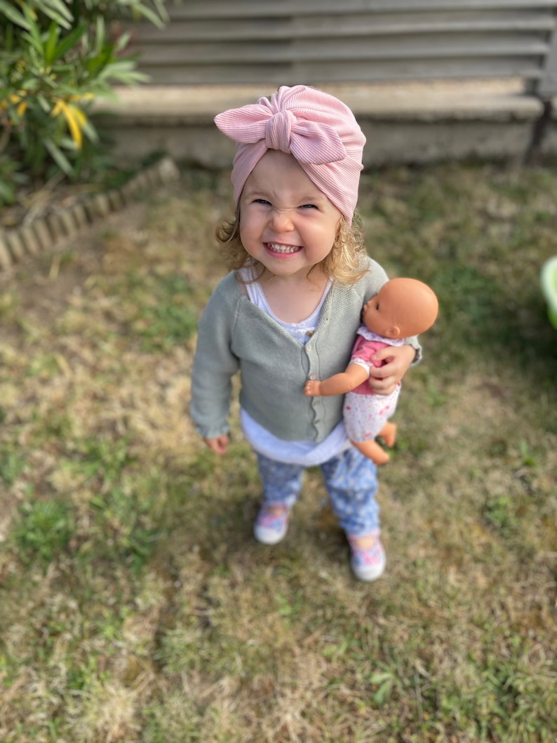 May include: A young child wearing a pink turban with a bow on top is holding a doll. The child is smiling and looking at the camera. The child is wearing a light green cardigan and blue jeans. The child is standing on a grassy surface.