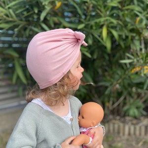 May include: A young child wearing a pink turban style hat holds a doll in their arms. The child is wearing a light green cardigan and the doll is wearing a pink and white dress.