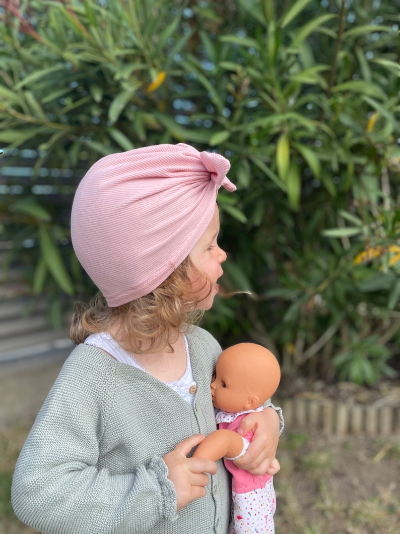 May include: A young child wearing a pink turban style hat holds a doll in their arms. The child is wearing a light green cardigan and the doll is wearing a pink and white dress.
