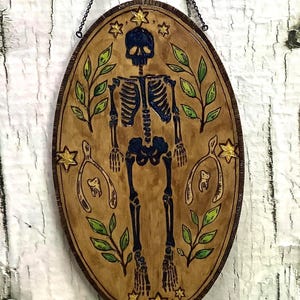 Puede incluir: Colgante de pared ovalado de madera con un diseño de esqueleto pintado a mano. El esqueleto es azul oscuro, rodeado de hojas verdes y estrellas amarillas. La obra de arte está suspendida por una cadena de metal.