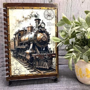 May include: A spiral-bound notebook with a detailed illustration of a vintage steam train. The cover has a brown leather-like texture with brass-coloured corner accents. The train is brown and gold, with white smoke billowing from the smokestack.