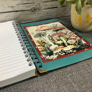 Puede incluir: Cuaderno de espiral abierto con p&aacute;ginas rayadas. La cubierta turquesa presenta una colorida ilustraci&oacute;n de setas. Las esquinas est&aacute;n protegidas con metal dorado. Una peque&ntilde;a planta en maceta est&aacute; en el fondo.