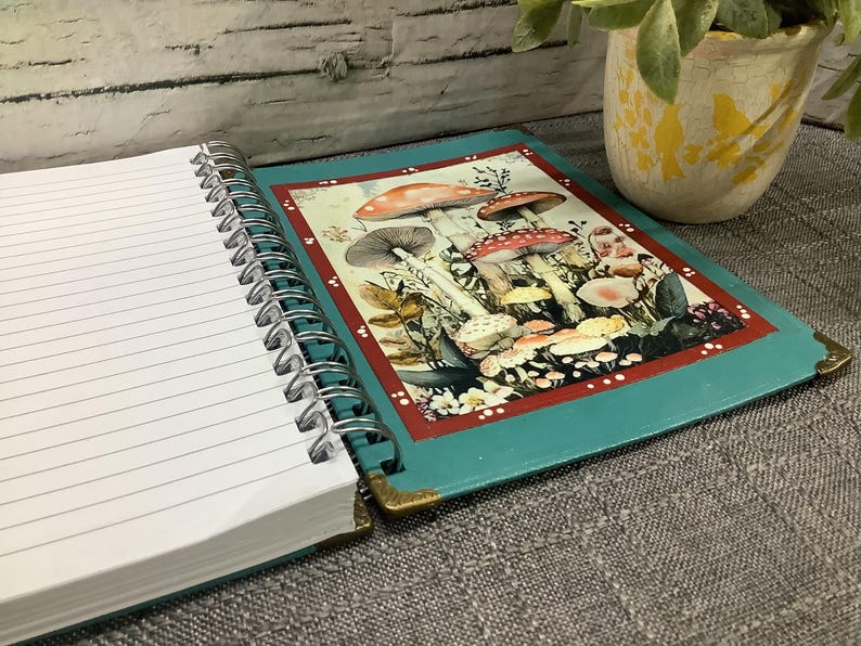 Puede incluir: Cuaderno de espiral abierto con p&aacute;ginas rayadas. La cubierta turquesa presenta una colorida ilustraci&oacute;n de setas. Las esquinas est&aacute;n protegidas con metal dorado. Una peque&ntilde;a planta en maceta est&aacute; en el fondo.