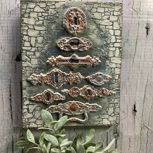 May include: A rustic wooden plaque with a stone-like texture features a collection of antique-style metal door hardware. The hardware is arranged in a pyramid shape, with the largest piece at the top and the smallest at the bottom. The hardware is painted in shades of copper and silver, giving it a vintage look.