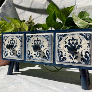 May include: A small, three-drawer wooden storage chest with a navy blue frame and legs. The drawers feature a distressed white and blue floral tile design and dark blue knobs. The chest is a decorative accent piece.