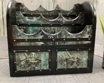 Puede incluir: Un organizador de madera negro con un acabado desgastado y detalles metálicos. Tiene cinco compartimentos y dos cajones con tiradores de latón. El organizador está decorado con un diseño de inspiración vintage.