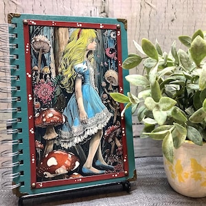 Puede incluir: Un cuaderno con espiral con una colorida ilustraci&oacute;n de Alicia en el Pa&iacute;s de las Maravillas. La cubierta tiene un borde turquesa y un marco interior rojo. La obra de arte representa a Alicia con un vestido azul, rodeada de setas y elementos del bosque.