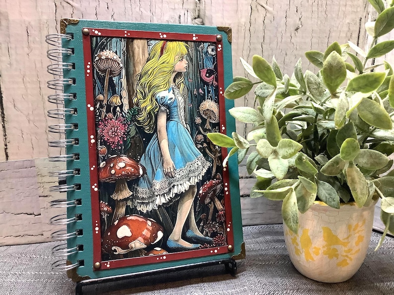 Puede incluir: Un cuaderno con espiral con una colorida ilustraci&oacute;n de Alicia en el Pa&iacute;s de las Maravillas. La cubierta tiene un borde turquesa y un marco interior rojo. La obra de arte representa a Alicia con un vestido azul, rodeada de setas y elementos del bosque.