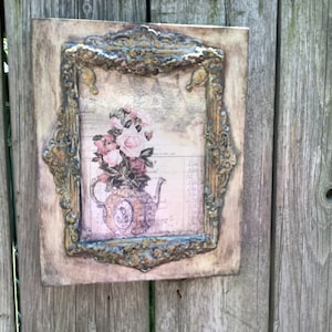 May include: A rectangular wooden plaque with a vintage-style floral print. The print features a teapot and pink roses within a decorative gold-colored frame. The frame has ornate detailing and is mounted on a weathered wooden background.
