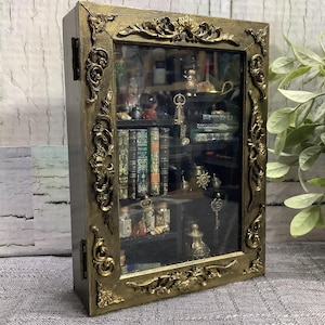 May include: A rectangular wall-mounted display cabinet with a dark gold, ornate, carved frame and a glass front door. The interior features multiple shelves containing miniature books, small glass bottles, and various metallic trinkets.