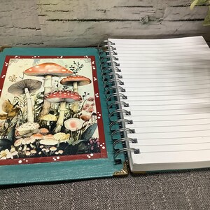 Puede incluir: Un cuaderno espiral abierto con una cubierta turquesa que presenta una ilustraci&oacute;n de hongos. El cuaderno tiene p&aacute;ginas rayadas y una encuadernaci&oacute;n en espiral plateada. El arte de los hongos tiene tapas rojas, naranjas y marrones.