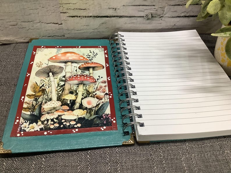 Puede incluir: Un cuaderno espiral abierto con una cubierta turquesa que presenta una ilustraci&oacute;n de hongos. El cuaderno tiene p&aacute;ginas rayadas y una encuadernaci&oacute;n en espiral plateada. El arte de los hongos tiene tapas rojas, naranjas y marrones.