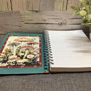 Puede incluir: Un cuaderno abierto con espiral con p&aacute;ginas rayadas junto a una revista turquesa con una ilustraci&oacute;n de hongos. La revista tiene un borde rojo y protectores de esquina dorados. Una peque&ntilde;a planta en maceta est&aacute; en el fondo.