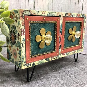 May include: A floral-patterned cabinet with two doors, each with a gold flower knob and a teal centre framed in red. The cabinet sits on black hairpin legs, adding a vintage touch. The cabinet's dimensions are approximately 30 cm in length.