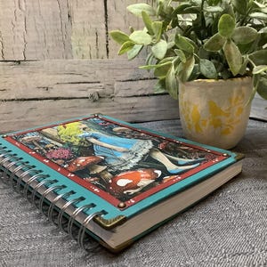 Puede incluir: Un cuaderno con espiral con una cubierta turquesa y una colorida ilustraci&oacute;n de Alicia en el pa&iacute;s de las maravillas. La portada presenta a Alicia con un vestido azul, setas y otros elementos caprichosos. El cuaderno tiene protectores de esquina de lat&oacute;n.