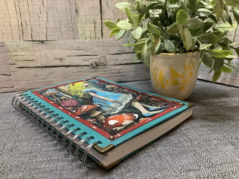 Puede incluir: Un cuaderno con espiral con una cubierta turquesa y una colorida ilustraci&oacute;n de Alicia en el pa&iacute;s de las maravillas. La portada presenta a Alicia con un vestido azul, setas y otros elementos caprichosos. El cuaderno tiene protectores de esquina de lat&oacute;n.