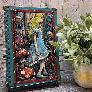 Puede incluir: Un cuaderno con espiral con una cubierta turquesa y una colorida ilustraci&oacute;n de Alicia en el Pa&iacute;s de las Maravillas. La obra de arte presenta a Alicia con un vestido azul rodeada de setas y flores. El cuaderno est&aacute; apoyado con un peque&ntilde;o soporte.