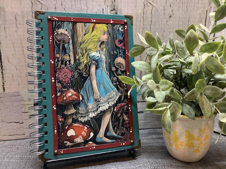 Puede incluir: Un cuaderno con espiral con una cubierta turquesa y una colorida ilustraci&oacute;n de Alicia en el Pa&iacute;s de las Maravillas. La obra de arte presenta a Alicia con un vestido azul rodeada de setas y flores. El cuaderno est&aacute; apoyado con un peque&ntilde;o soporte.