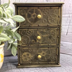 May include: A three-drawer steampunk-style chest with a bronze finish. The drawers are decorated with gear and cog designs and have round, gold-colored knobs. The chest is set against a rustic, white-washed wood background.