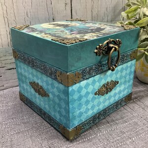 Op de afbeelding: Vierkante, turquoise decoratieve doos met een scharnierend deksel. De doos heeft een ruitpatroon op de zijkanten en sierlijke metalen hoekaccenten. Het deksel heeft een gedrukte afbeelding en een metalen handvat.