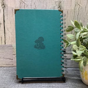Puede incluir: Un cuaderno en espiral turquesa con un dise&ntilde;o de setas. El cuaderno tiene protectores de esquina dorados y se muestra sobre un soporte negro. Una peque&ntilde;a planta en maceta con hojas verdes est&aacute; en el fondo.