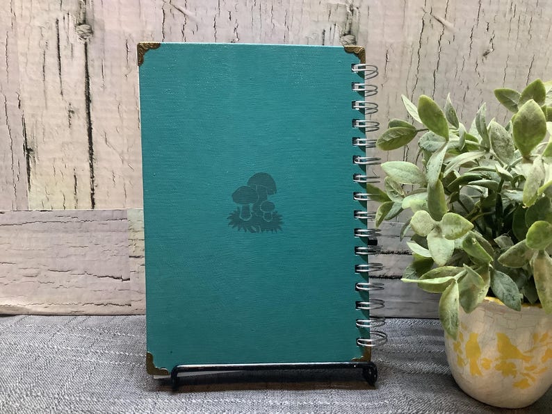 Puede incluir: Un cuaderno en espiral turquesa con un dise&ntilde;o de setas. El cuaderno tiene protectores de esquina dorados y se muestra sobre un soporte negro. Una peque&ntilde;a planta en maceta con hojas verdes est&aacute; en el fondo.