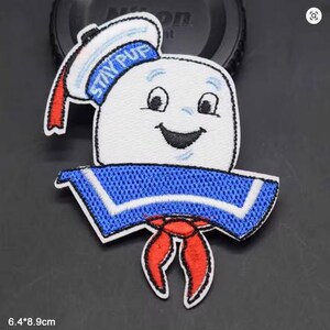 Könnte beinhalten: Ein gestickter Aufnäher mit dem Charakter Stay Puft Marshmallow Man. Das Design zeigt eine weiße Marshmallow-Figur mit lächelndem Gesicht, die einen blauen Matrosenkragen, ein rotes Halstuch und eine weiße Matrosenmütze mit dem Text STAY PUFT trägt.