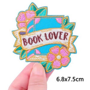 Könnte beinhalten: Ein gestickter Aufnäher mit einem hellblauen Buch, umgeben von rosa Blumen und grünen Blättern. Ein weißes Banner in der Mitte zeigt den Text BOOK LOVER in braunen Buchstaben. Der Aufnäher misst insgesamt 6,8 mal 7,5 Zentimeter.