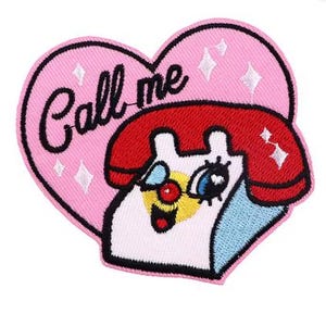 May include: An embroidered patch in the shape of a pink heart featuring a red and white rotary telephone with a smiling face. The text Call me is stitched in black cursive script above the telephone, accented by small white sparkling star icons.