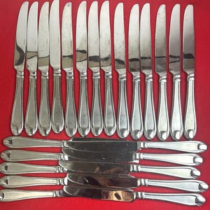 May include: A collection of stainless steel knives with a reflective finish. The knives are arranged in rows on a red surface. The handles have a decorative, elongated design. The blades are curved and appear sharp. The image showcases a set of cutlery.
