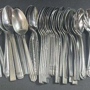 Puede incluir: Una gran colección de cubiertos de acero inoxidable en tono plateado que incluye varias cucharas, una cuchara de servir con borde festoneado y dos cuchillos de mantequilla ornamentados con mangos florales detallados alineados sobre una superficie oscura.