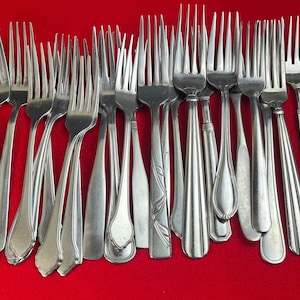 May include: A collection of stainless steel forks with various handle designs. The forks are arranged on a red surface, showcasing different styles, including smooth, ridged, and floral patterns. The tines are uniform, and the overall presentation suggests a set of silverware.
