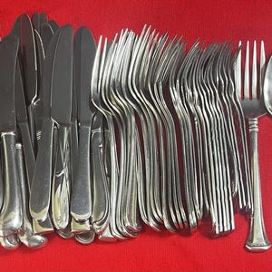 Puede incluir: Una gran colección de cubiertos de acero inoxidable en tono plateado que incluye varios cuchillos, tenedores y cucharas. El conjunto presenta un diseño de mango simple y liso con un acabado pulido, dispuesto en fila sobre un fondo rojo.