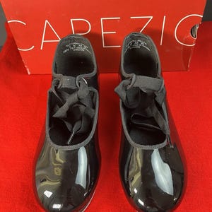 Puede incluir: Un par de zapatos de claqué de charol negro con cordones de cinta, sobre una superficie roja. Los zapatos están frente a una caja roja con el nombre de la marca "Capezio" impreso.