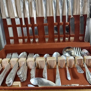 May include: A vintage silver cutlery set presented in a wooden case with a red velvet lining. The set includes knives, forks, and spoons, each with decorative handle detailing. The arrangement suggests a complete service for multiple place settings.