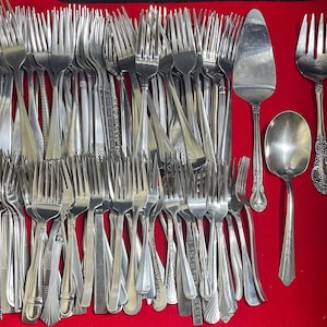 May include: A collection of various silver forks, a pie server, and a spoon on a red surface. The forks have different handle designs, and the pie server has a wide, flat blade. The spoon has a rounded bowl.