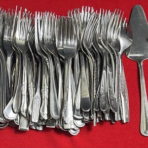 May include: A collection of silver forks, a cake server, a soup spoon, and a serving fork on a red surface. The forks have various handle designs. The cake server and spoon have simple, elegant designs. The serving fork has a decorative handle.